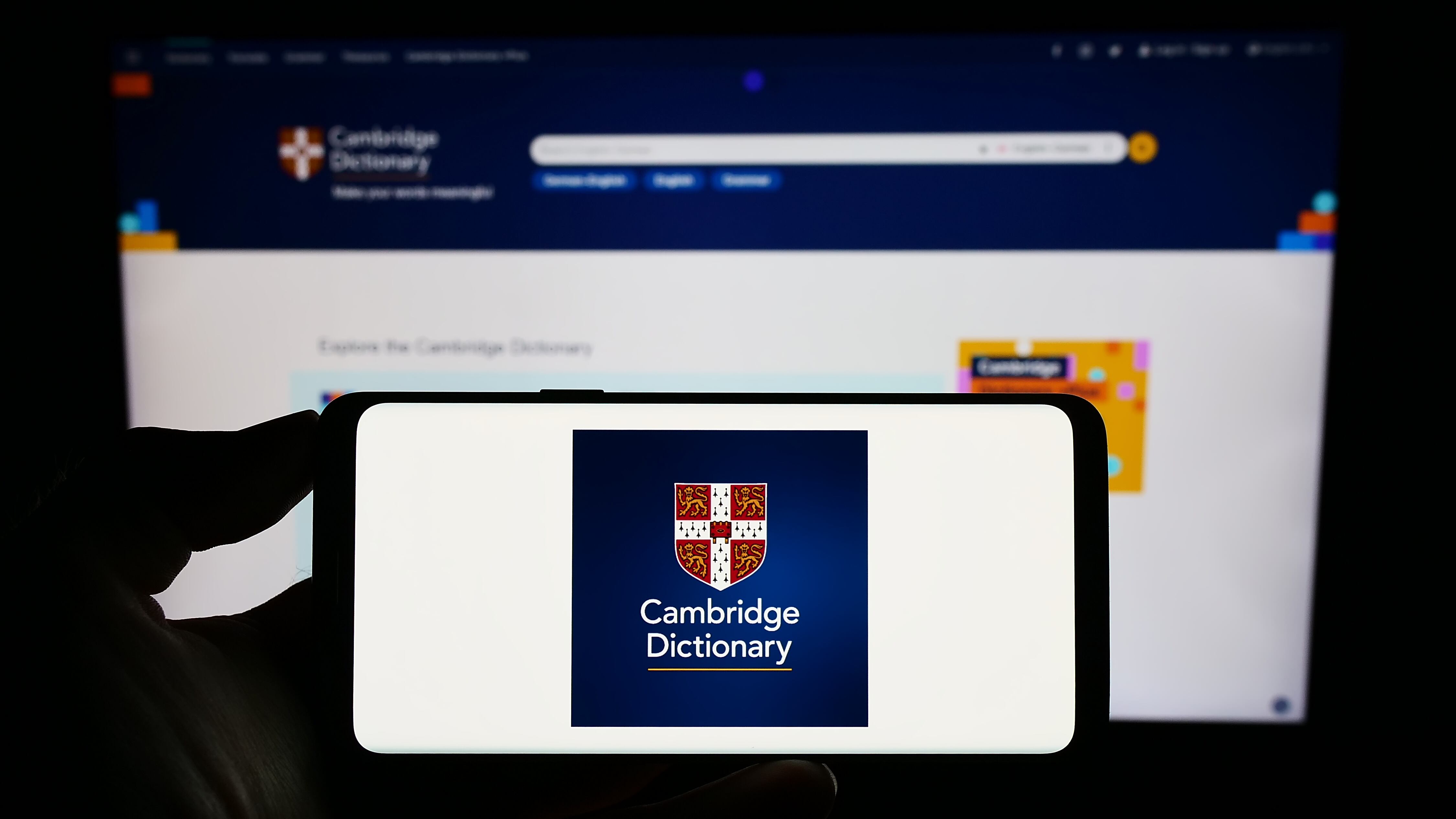 Person holding smartphone with logo of Cambridge Dictionary on screen in front of website. Focus on phone display.